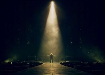 Figura solitária no centro de um palco iluminado por um único spotlight, simbolizando a solidão e a impotência retratadas em Bad do U2.