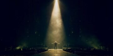 Figura solitária no centro de um palco iluminado por um único spotlight, simbolizando a solidão e a impotência retratadas em Bad do U2.