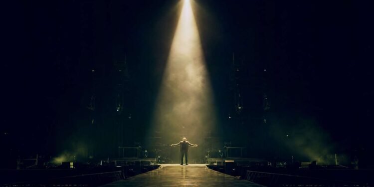 Figura solitária no centro de um palco iluminado por um único spotlight, simbolizando a solidão e a impotência retratadas em Bad do U2.