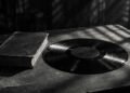 Disco de vinil escuro sobre uma mesa de arquivo, com um exemplar antigo de livro ao lado, em preto e branco