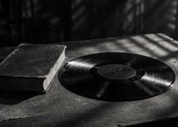 Disco de vinil escuro sobre uma mesa de arquivo, com um exemplar antigo de livro ao lado, em preto e branco