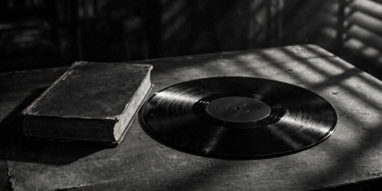 Disco de vinil escuro sobre uma mesa de arquivo, com um exemplar antigo de livro ao lado, em preto e branco