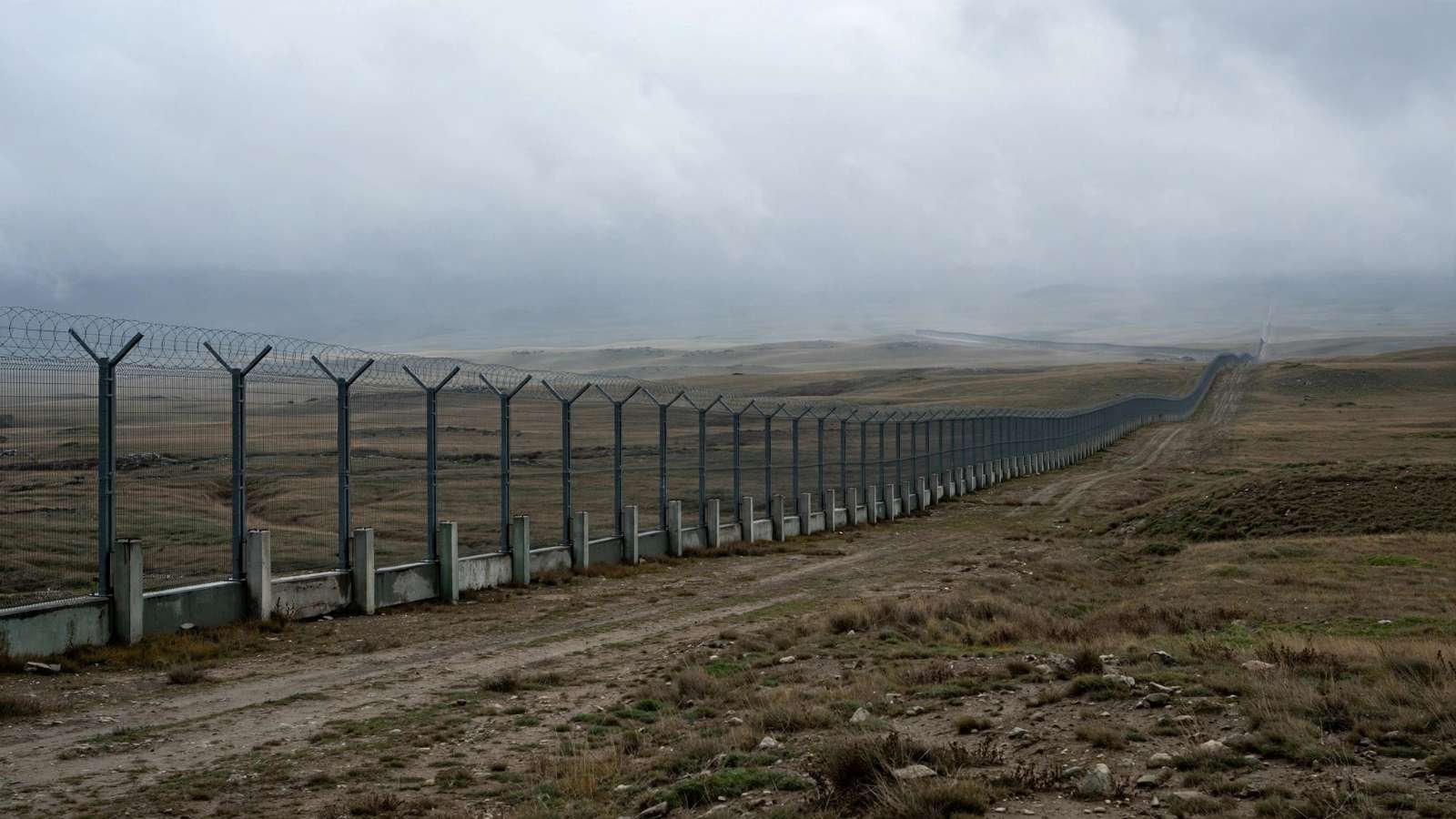 Fronteira sem nome Paisagem de fronteira fortificada, com cerca atravessando terreno aberto sob céu nublado, sem pessoas